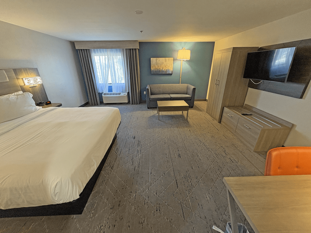 A spacious guest room at the Holiday Inn Express Tucson Airport featuring a king bed, a sofa, a desk, a large TV, and windows with curtains.