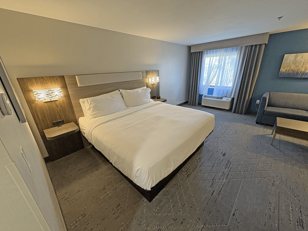 A spacious hotel guest room featuring a king-sized bed, modern furnishings, and soft lighting. There is a seating area with a couch, a window with curtains, and a desk beside the bed.