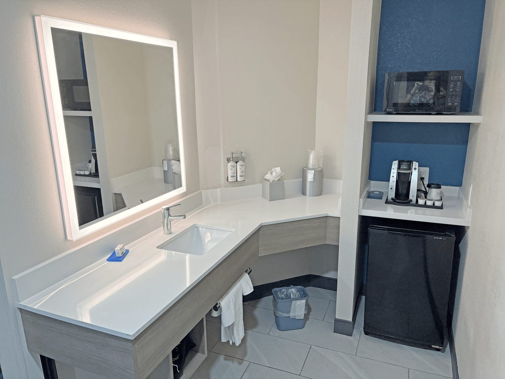 A modern hotel room bathroom featuring a large illuminated mirror, a white countertop with a sink, and convenient amenities including a coffee maker and microwave on a shelf.