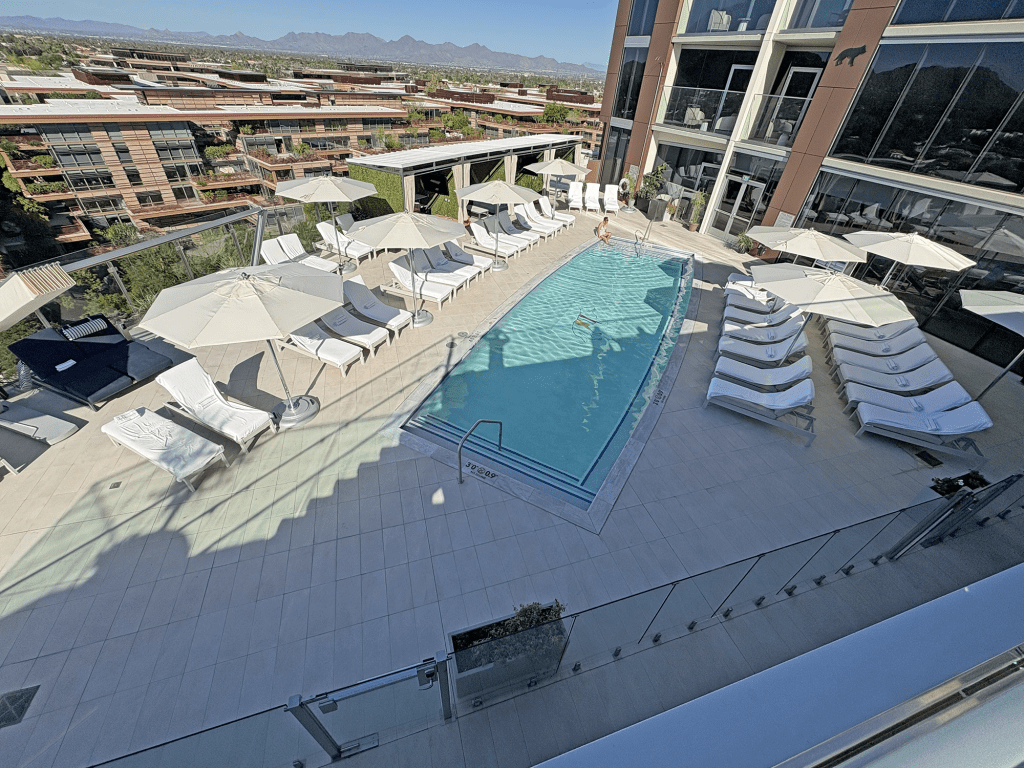 Aerial view of a modern hotel pool area featuring lounge chairs and umbrellas, surrounded by greenery and buildings with mountain views in the background.